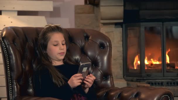 Маленька дівчинка проводить дозвілля зі смартфоном біля каміна — стокове відео