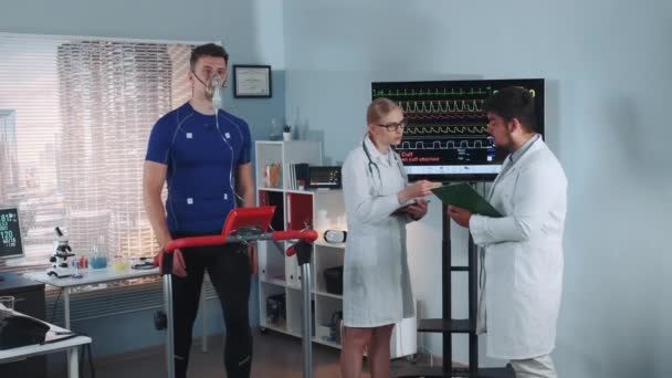 Multiracial scientists examining the athletes medical records while he stress testing in lab — Stock Video