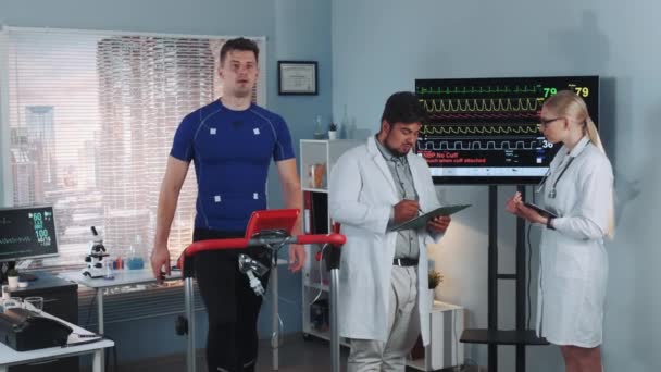 Dois médicos de raça mista discutindo dados de ECG de atleta fazendo teste de esteira no laboratório de esportes — Vídeo de Stock