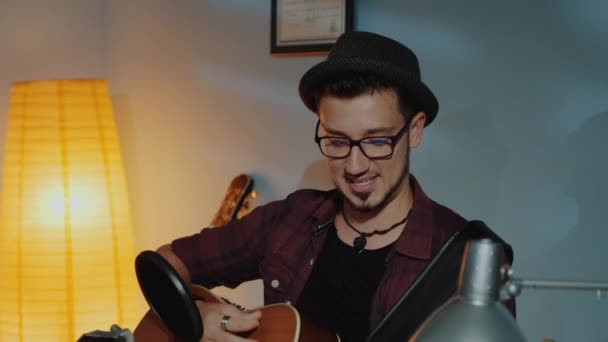 Primer plano de tipo barbudo en sombrero emocionalmente cantando en el micrófono y tocando la guitarra en el estudio en casa — Vídeos de Stock
