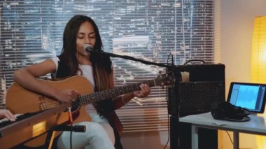 Ev stüdyosundaki grup provasında gitar çalan ve mikrofona şarkı söyleyen melez bir genç bayan.