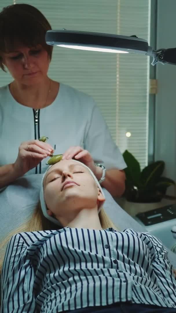 Hermosa mujer recibiendo masaje facial con rodillo de jade en el salón de belleza con poca luz alrededor — Vídeos de Stock