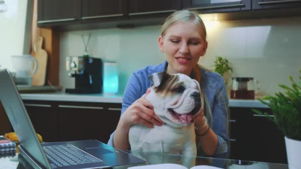 Vrolijke jonge vrouw met bulldog puppy zittend voor laptop in de keuken — Stockvideo