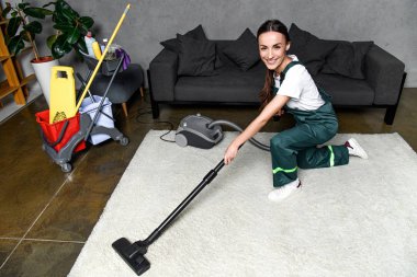 high angle view of happy young female cleaner using vacuum cleaner and smiling at camera while cleaning white carpet clipart