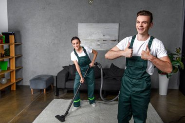 happy young cleaning company workers using vacuum cleaner and showing thumbs up clipart