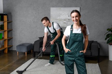 young cleaning company workers using vacuum cleaner and smiling at camera clipart