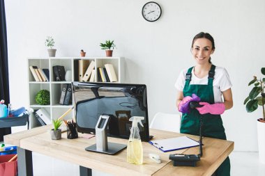 beautiful young professional cleaner smiling at camera while cleaning modern office clipart