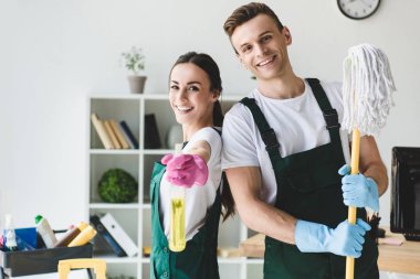 office temizlerken kameraya gülümseyen paspas ve sprey şişeyle mutlu genç temizleyiciler