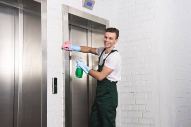 handsome young cleaner washing elevator and smiling at camera  clipart