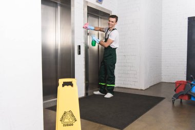 handsome young janitor cleaning elevator and smiling at camera  clipart