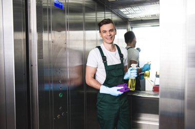 handsome young janitor holding spray bottle with detergent and rag, smiling at camera in elevator clipart