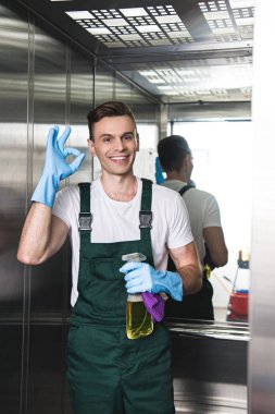 handsome young janitor holding spray bottle with detergent and rag, smiling at camera and showing ok sign in elevator clipart