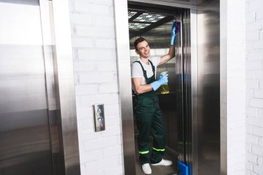 handsome young janitor cleaning elevator and smiling at camera   clipart