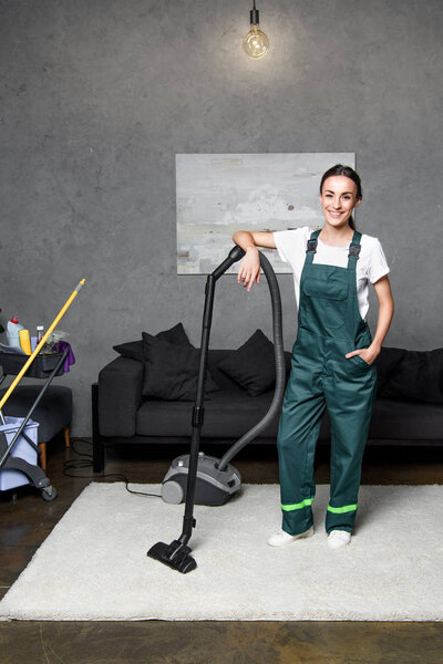 happy young female cleaning company worker leaning at vacuum cleaner and smiling at camera 