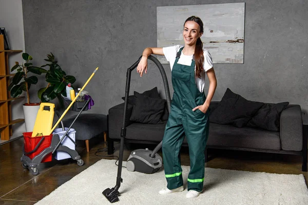 年轻女性清洁公司工人靠在真空吸尘器和微笑的相机 — 图库照片