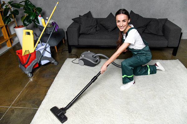 high angle view of happy young female cleaner using vacuum cleaner and smiling at camera while cleaning white carpet