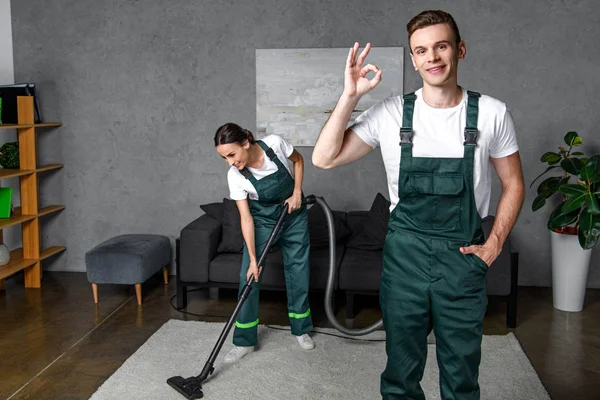 Tersenyum Muda Pekerja Pembersih Menggunakan Vacuum Cleaner Dan Menunjukkan Tanda — Stok Foto