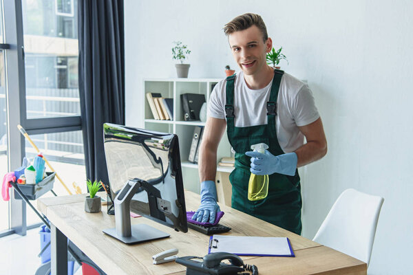 handsome professional cleaner in rubber gloves holding detergent spray and smiling at camera in office 