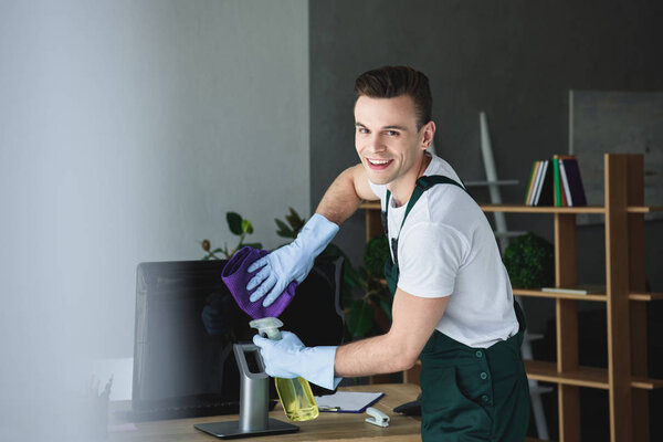 handsome young professional cleaner smiling at camera while cleaning computer monitor in office