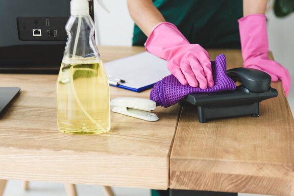 cropped shot of woman in rubber gloves cleaning telephone on table in office