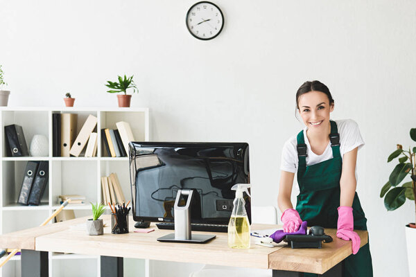 beautiful young cleaner smiling at camera while cleaning table in modern office