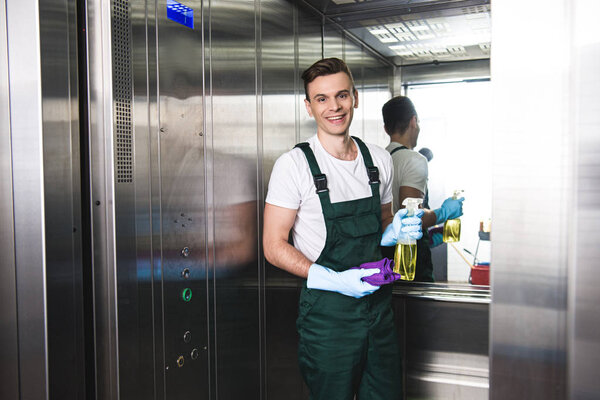 handsome young janitor holding spray bottle with detergent and rag, smiling at camera in elevator