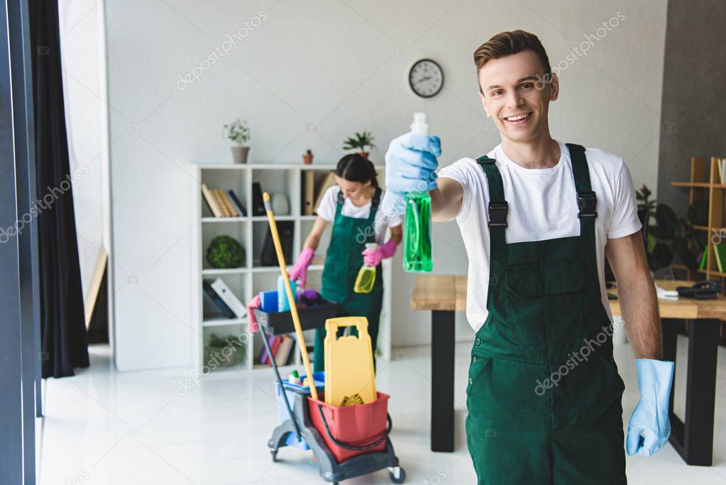 young male cleaner in rubber gloves holding spray bottle with detergent and smiling at camera