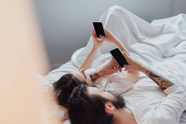 young couple lying in bed and using smartphones with blank screen