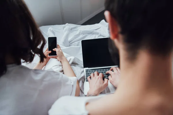 空白の画面を自宅のパソコンとスマート フォンを使用している間ベッドで横になっているカップルの背面図 — ストック写真