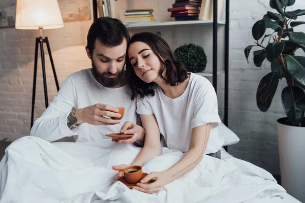 朝のコーヒーを飲みながらベッドに横たわっている美しい若いカップル — ストック写真