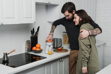 beautiful young couple making orange juice and hugging during breakfast in kitchen clipart
