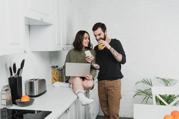man drinking juice and using smartphone while smiling woman using laptop during breakfast in kitchen