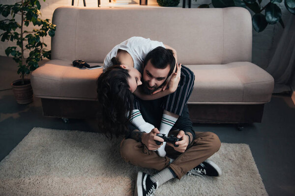 женщина обнимает и целует мужчину, играя в видеоигру с джойстиком в гостиной

