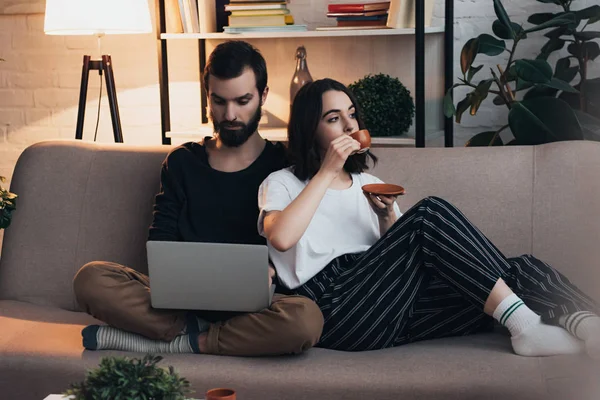 ソファに座って 居間でノート パソコンを使用している人 コーヒーを飲む女性 — ストック写真