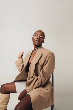 bej ceket ve kolye gri izole sandalyede oturan güzel afrikalı amerikalı kadın