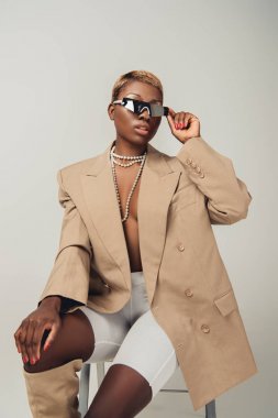 güneş gözlüğü ve gri sandalyede oturan zarif bej ceket şık afrikalı amerikalı kadın