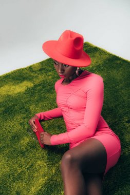 Pembe elbiseli ve şapkalı genç Afro-Amerikan manken yeşil çimlerin üzerinde çantasıyla poz veriyor.