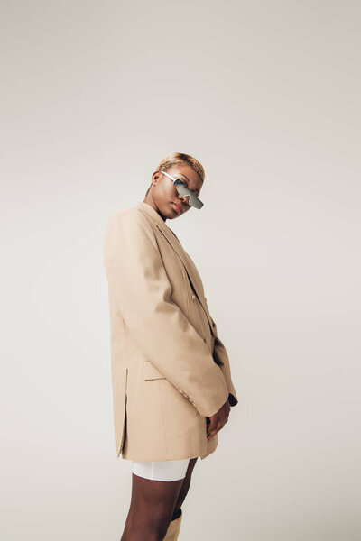 african american girl posing in sunglasses and beige jacket isolated on grey