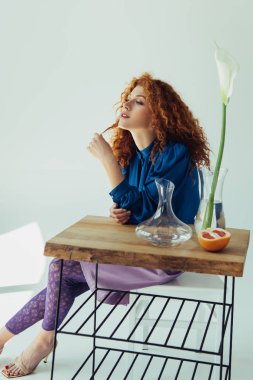 stylish redhead girl posing at table with calla flower and vases on grey clipart