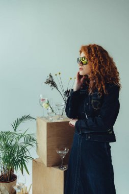 trendy redhead girl posing near wooden boxes, glasses and plants isolated on grey clipart