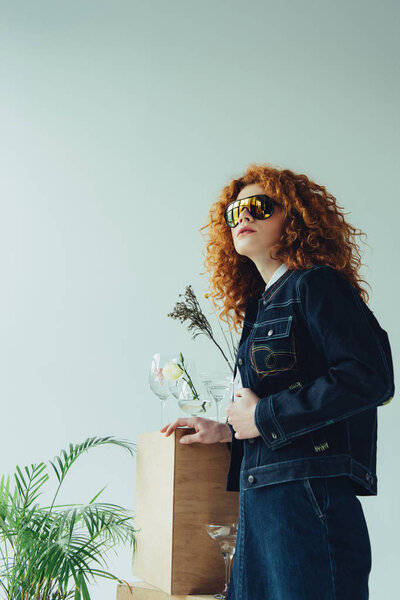 stylish redhead girl in sunglasses posing near wooden box and plants on grey