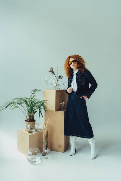 stylish redhead girl posing near wooden boxes, glasses and plants on grey