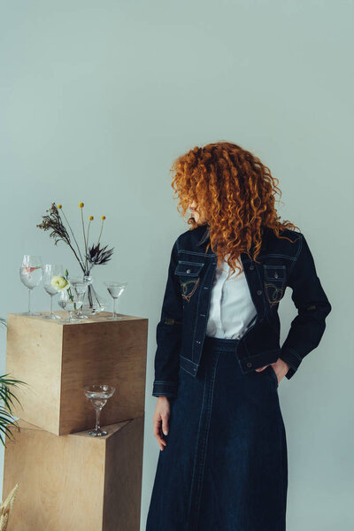 stylish redhead girl posing near wooden boxes, glasses and plants isolated on grey