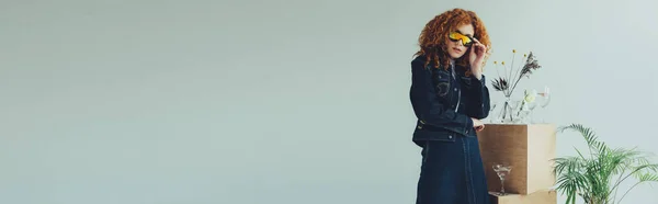 stock image panoramic shot of stylish redhead girl posing near glasses and plants isolated on grey with copy space