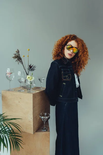 时髦的红发女孩在太阳镜上摆姿势附近的木箱 眼镜和植物的灰色 — 图库照片