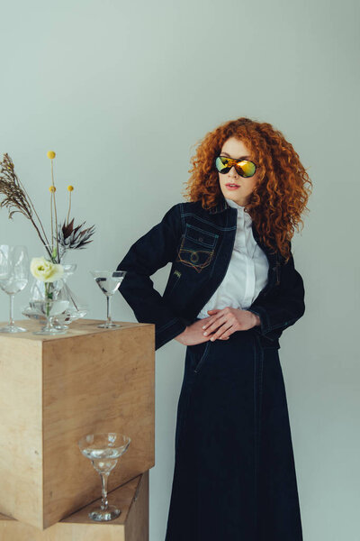 trendy redhead girl posing near wooden boxes, glasses and plants isolated on grey