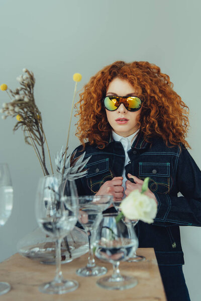 stylish redhead woman in sunglasses posing near glasses and dried flowers isolated on grey