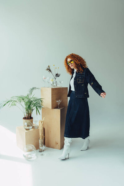 trendy redhead girl posing near wooden boxes, glasses and plants on grey