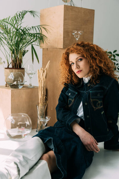 trendy redhead girl sitting and posing near wooden boxes, glasses and plants on grey