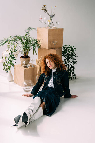stylish redhead girl posing near wooden boxes, glasses and plants on grey with sunlight